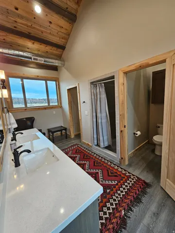 Double vanity with a light-toned countertop and matte black fixtures, exposed wood plank vaulted ceiling, and a tiled shower