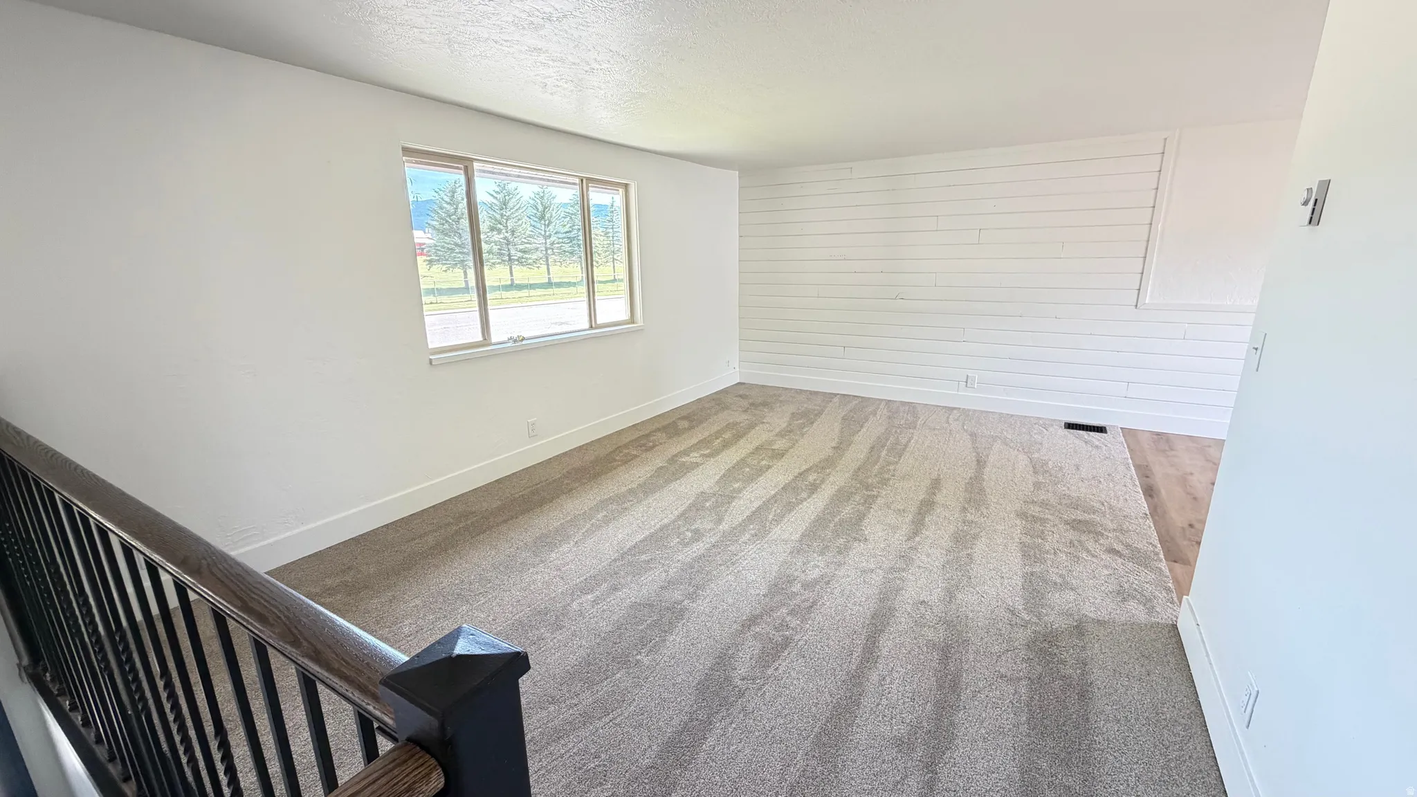 Open concept living area featuring a large window, shiplap accent wall, neutral carpeting, and a dark wood banister