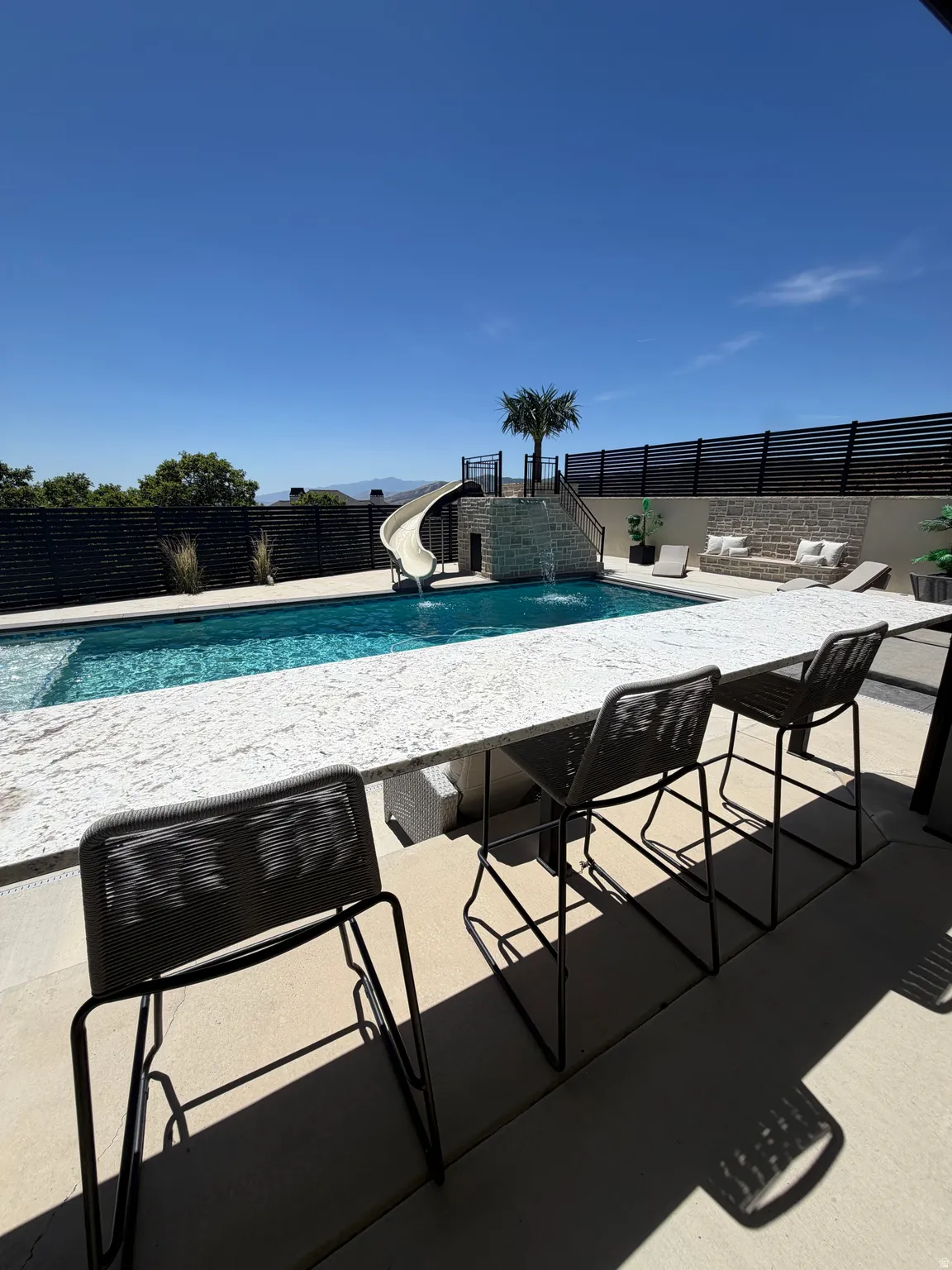 Outdoor entertaining space featuring a built-in bar, pool with water feature and slide, stone retaining walls, and privacy fencing