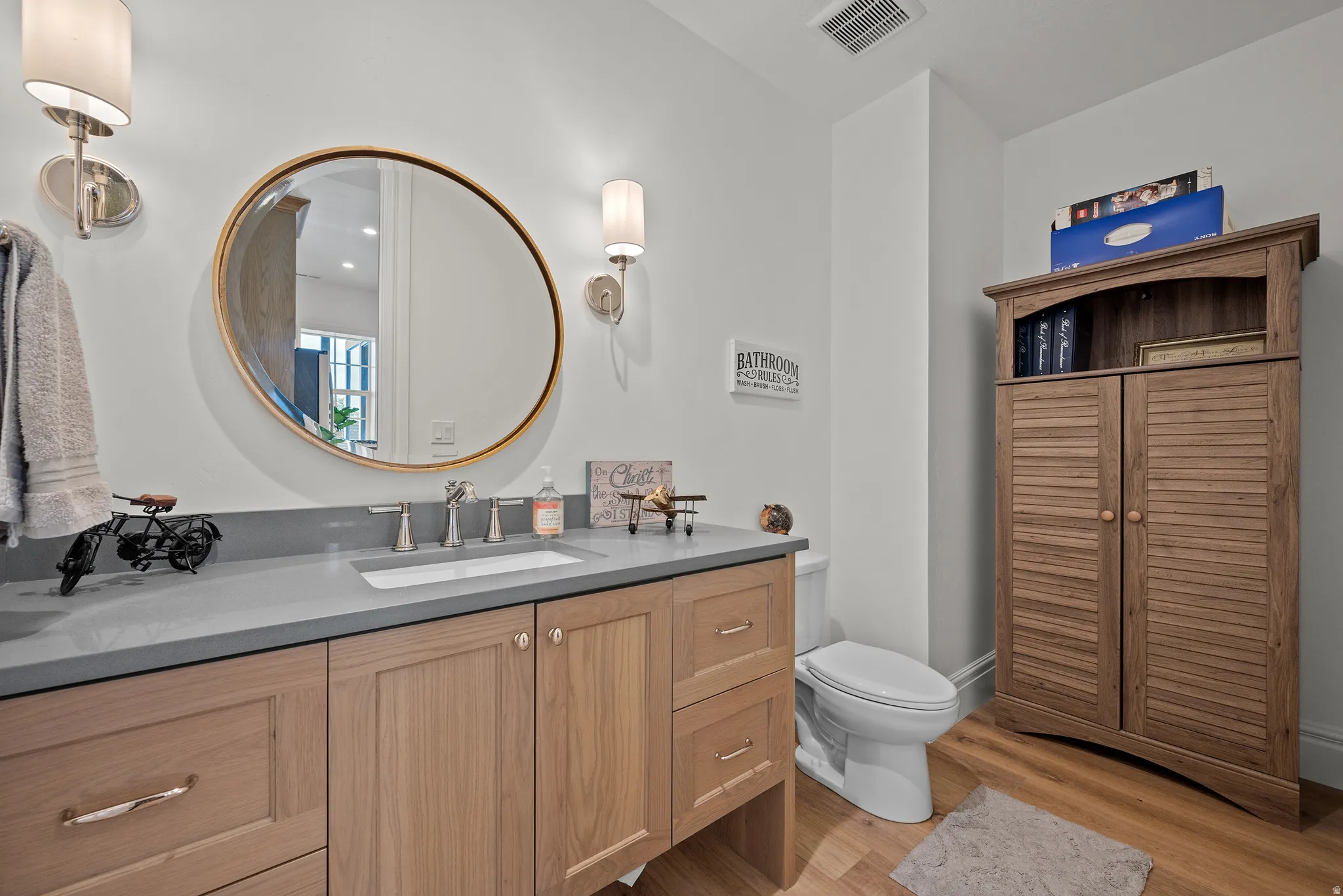 Bathroom featuring a light wood-finish vanity with a built-in sink and gray countertop, complemented by a round framed mirror