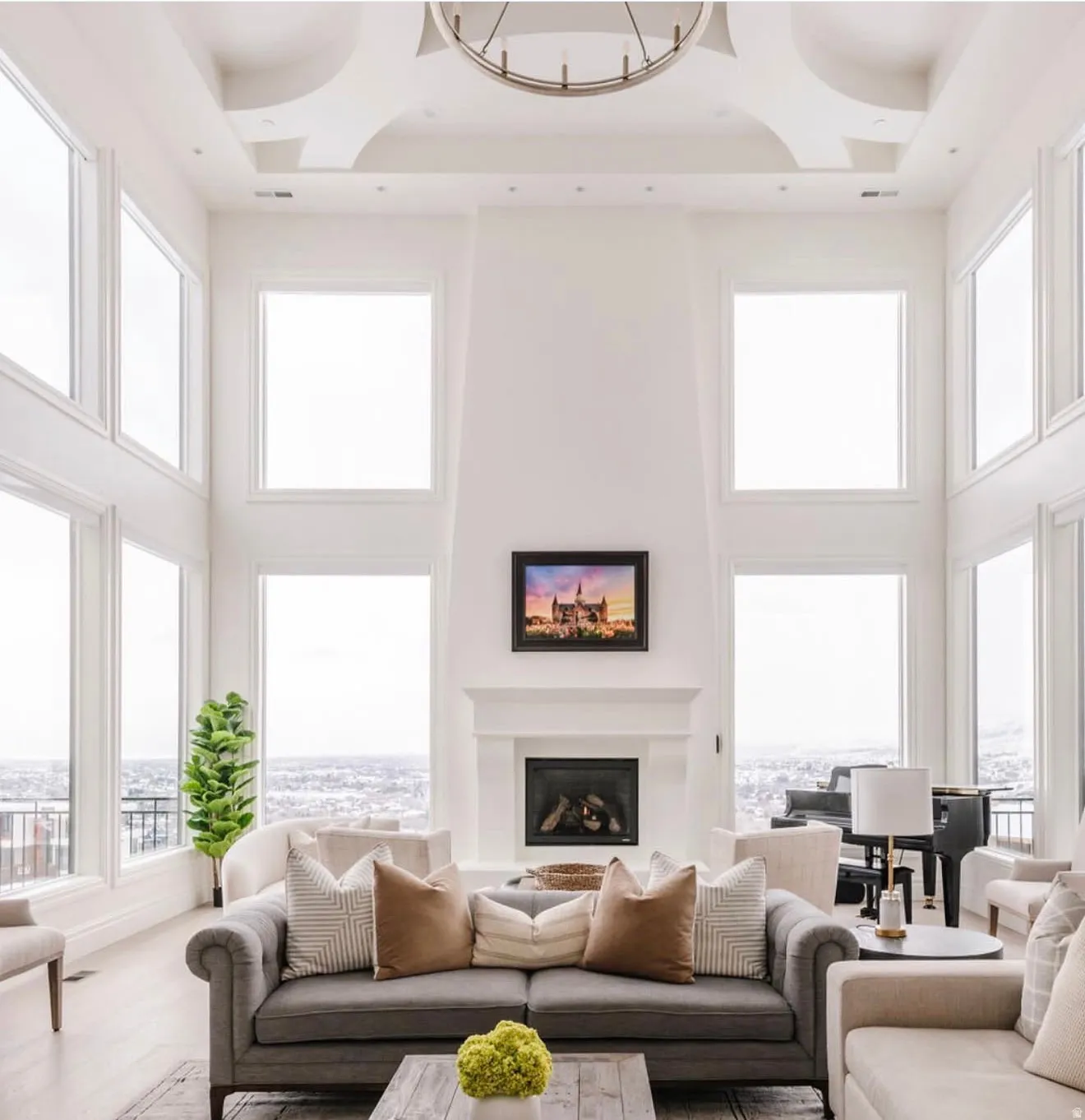 Spacious living area featuring a vaulted ceiling with a circular chandelier, a grand fireplace with a white mantel, and extensive windows showcasing distant scenery
