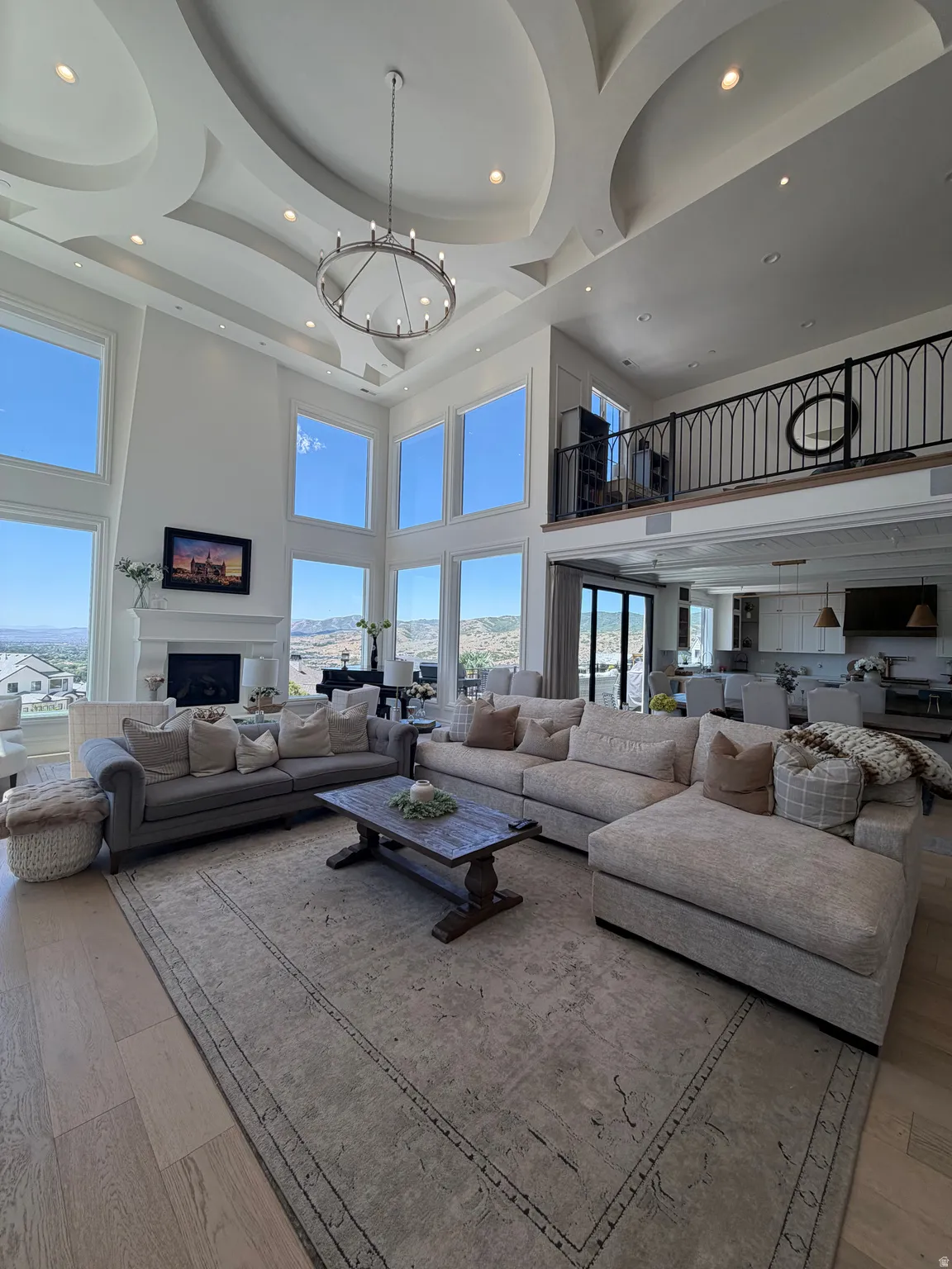 Two-story living area featuring a vaulted ceiling with decorative insets and recessed lighting, light wood-finish flooring, a contemporary chandelier, and a second-level balcony with an ornate black metal railing