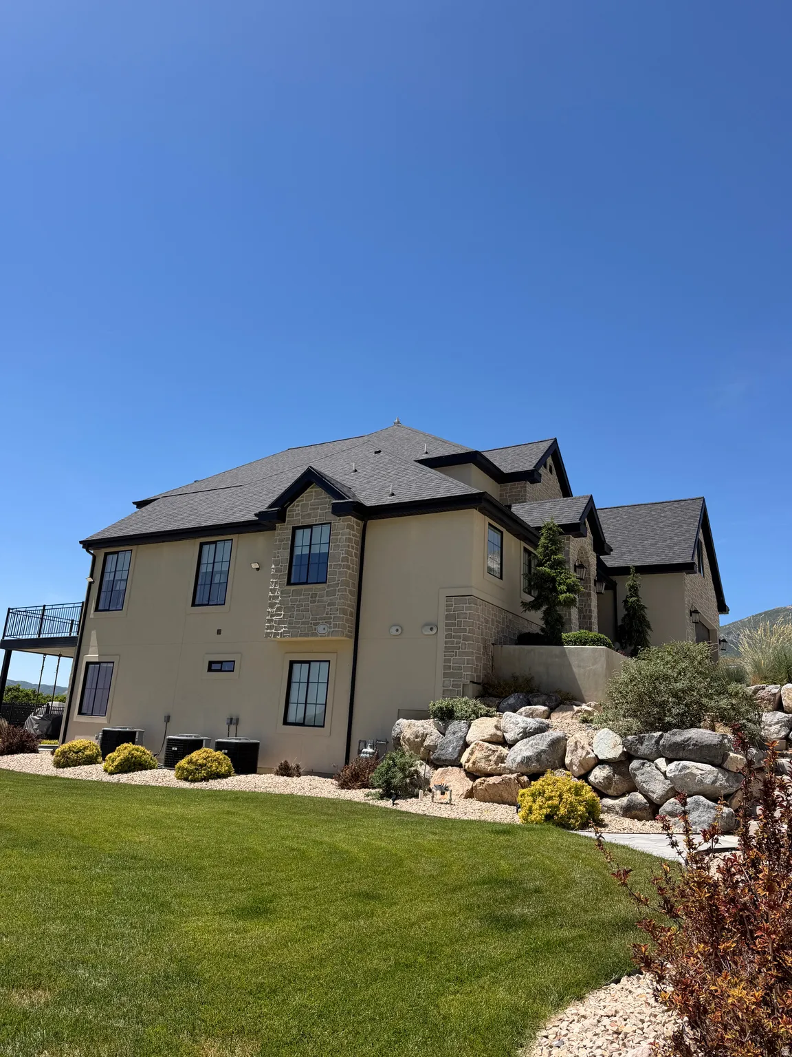Expansive rear exterior featuring stucco and stone veneer accents, a multi-level roofline with dark shingles, a raised metal deck, and a landscaped rock retaining wall with mature plantings