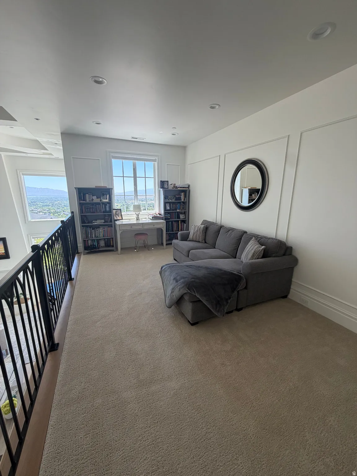 Open loft area featuring neutral-toned carpeting, ample natural light from multiple windows, recessed lighting, decorative wall paneling, and a black wrought iron railing