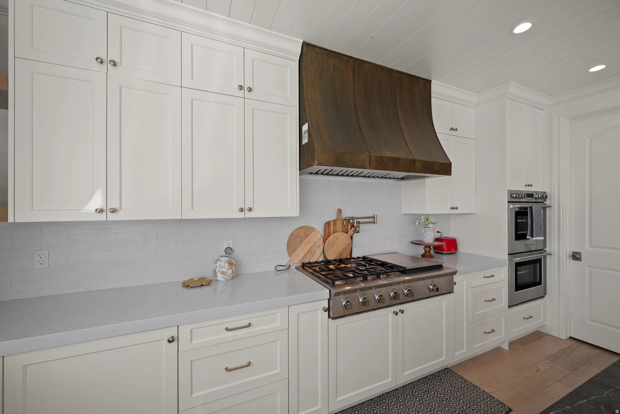 Kitchen featuring white shaker cabinetry, stainless steel gas range with griddle, custom dark metal range hood, light-toned countertops, and subway tile backsplash