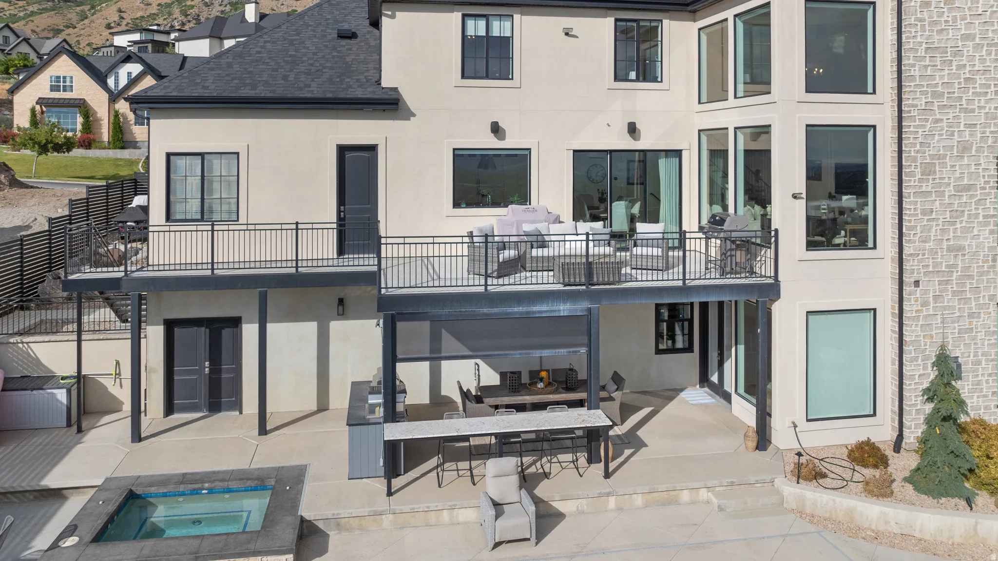 Multi-level stucco and stone exterior featuring a deck with black metal railing, lower patio with built-in outdoor kitchen, and an in-ground spa