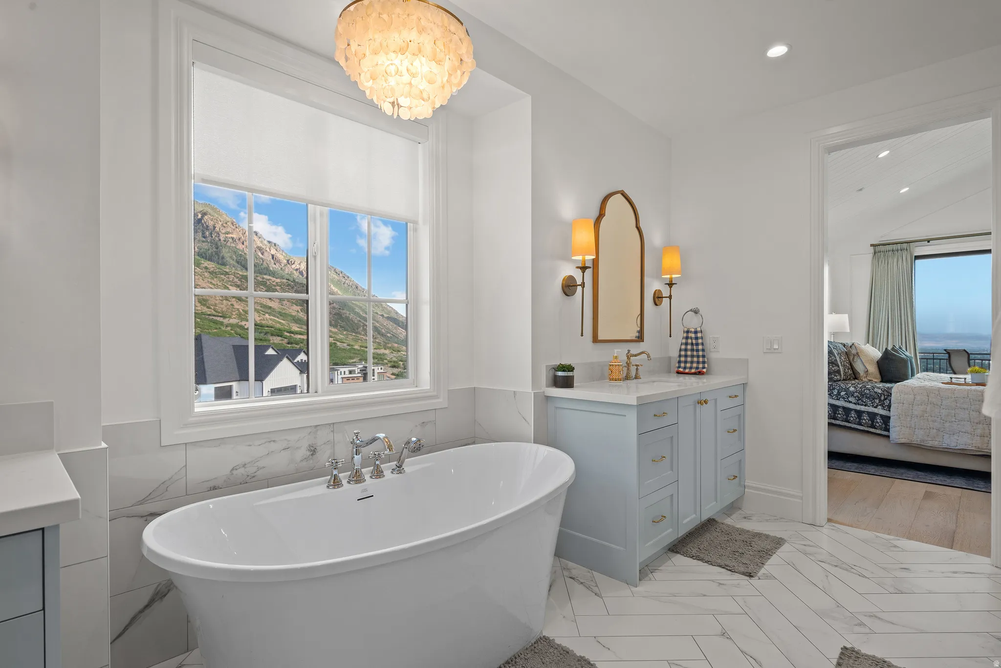 Bathroom featuring a freestanding soaking tub with deck-mounted fixtures, a window with mountain scenery, and a vanity with a light blue finish, a light-colored countertop, and a decorative mirror flanked by sconces