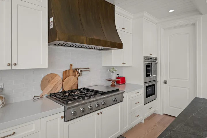 Gourmet kitchen featuring a substantial brass range hood, stainless steel gas cooktop, double wall ovens, white shaker cabinetry, and light-toned countertops