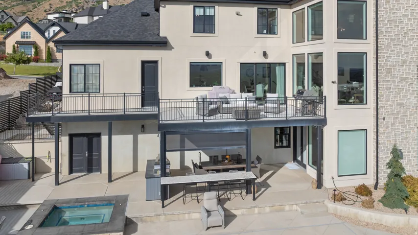 Multi-level stucco and stone exterior featuring a deck with black metal railing, lower patio with built-in outdoor kitchen, and an in-ground spa