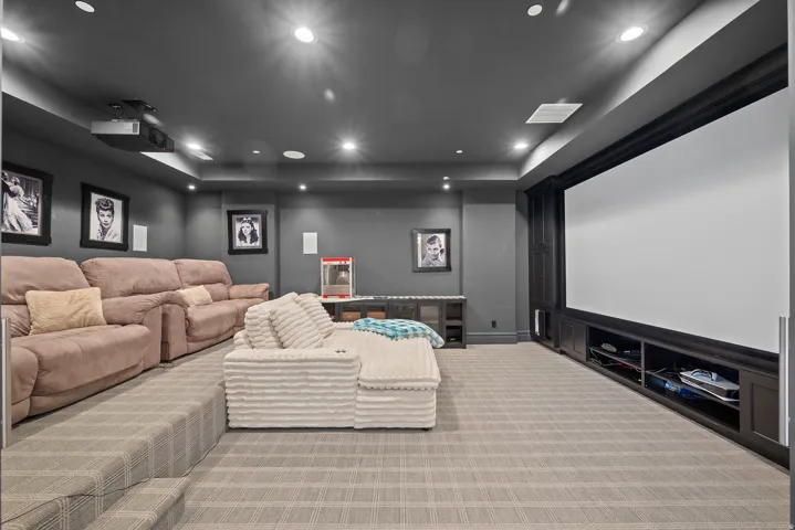 Dedicated media room featuring a large projection screen, tiered carpeted seating area, recessed ceiling lighting, and built-in dark wood cabinetry with open shelving