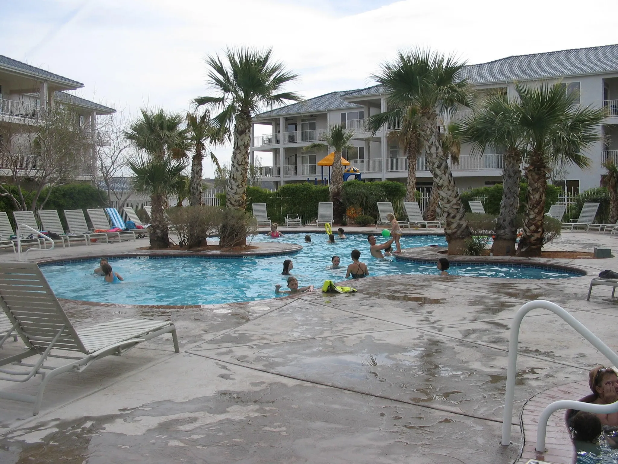 Expansive outdoor pool area featuring a structured concrete deck surround, integrated palm trees, and multiple chaise lounge chairs