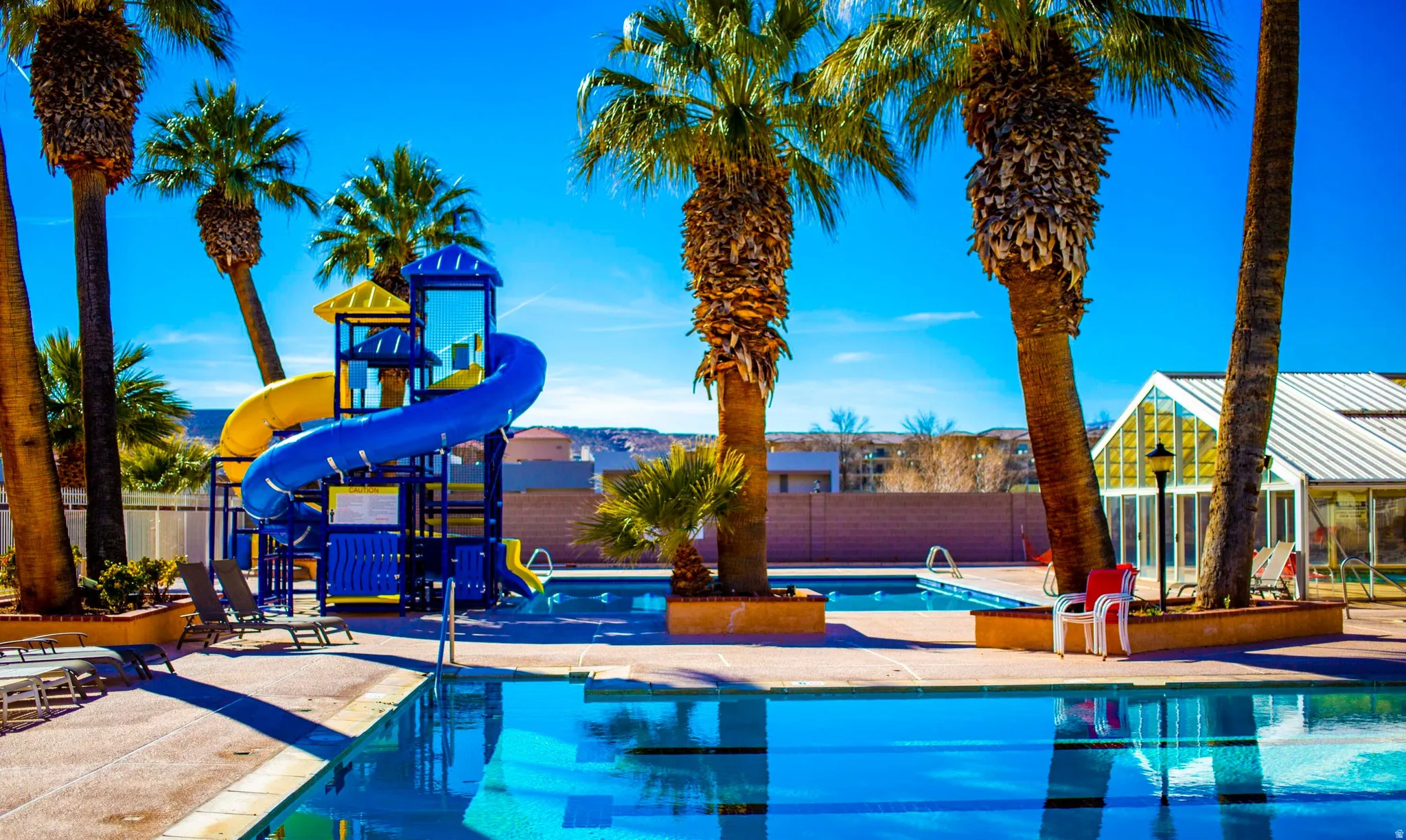 Outdoor swimming pool area featuring a prominent blue and yellow water slide, multiple mature palm trees, and a surrounding paved deck