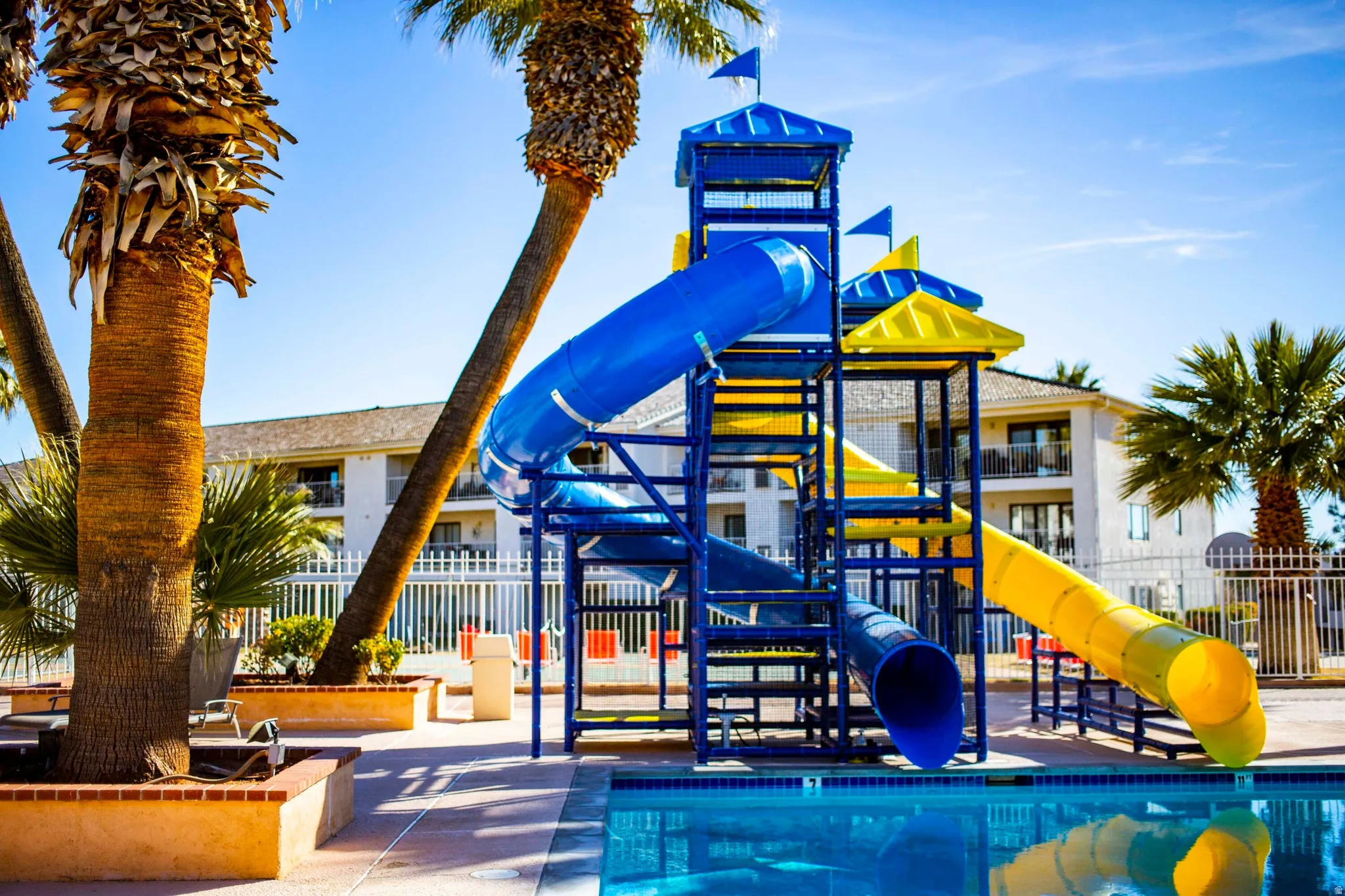 Aqua play structure with blue and yellow slides, surrounded by palm trees and concrete decking, beside a clear blue swimming pool