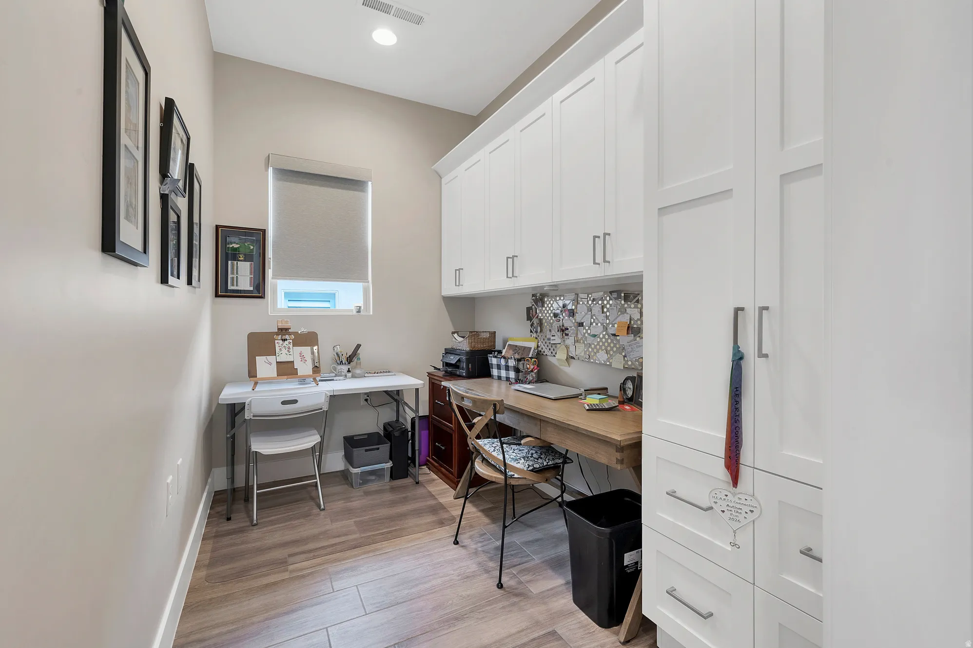 Dedicated workspace featuring extensive white cabinetry, a built-in wood-finish desk, wood-finish flooring, a window with a roller shade, and recessed lighting