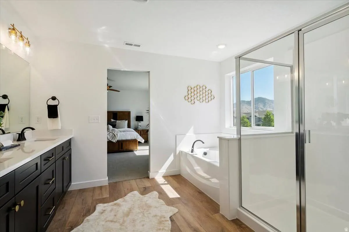Bathroom featuring a dual vanity with dark cabinetry, integrated soaking tub, separate glass-enclosed shower, wood-finish flooring, and a window with mountain scenery