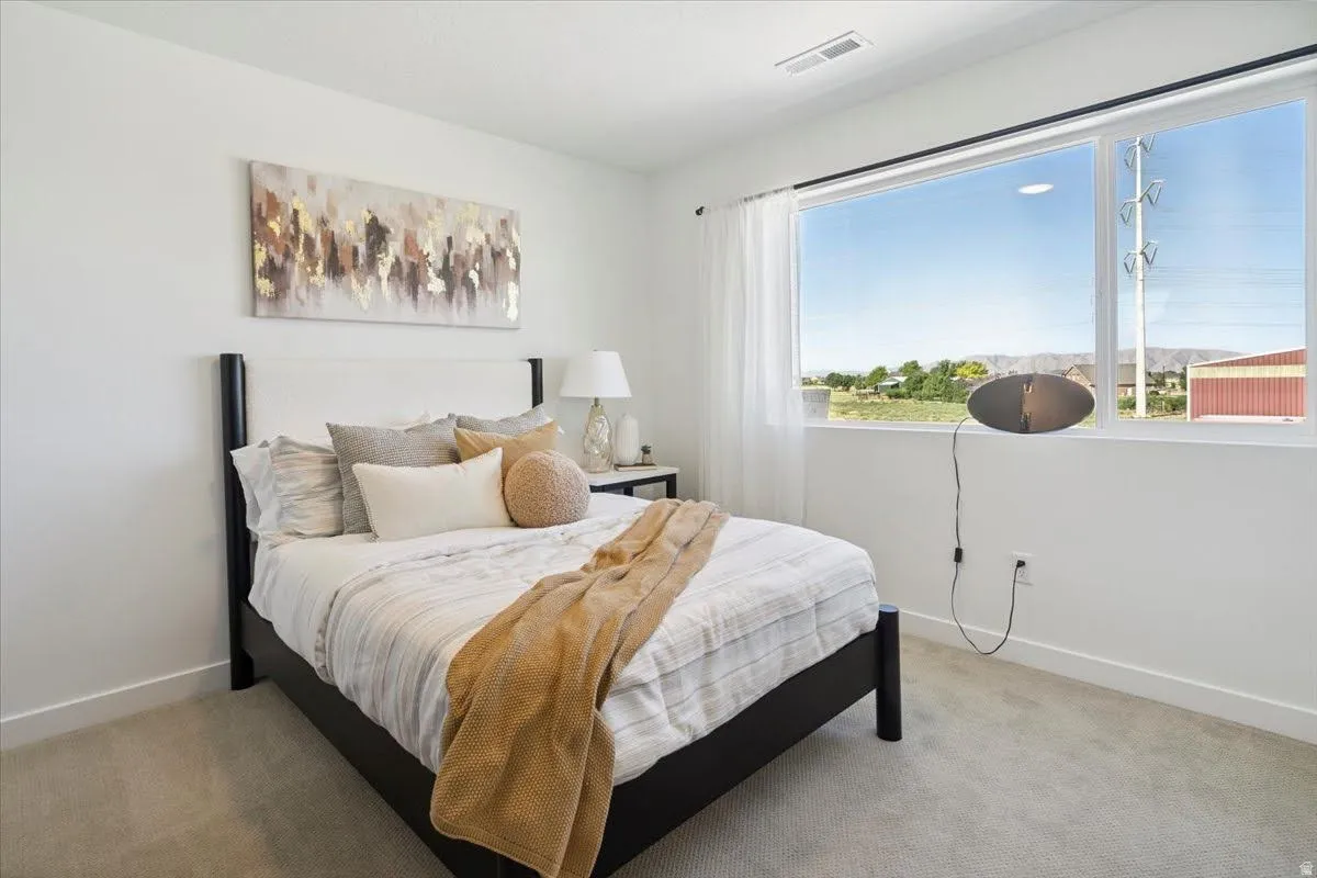 Bright bedroom featuring a large picture window, light-colored carpeting, white walls, and a recessed ceiling vent