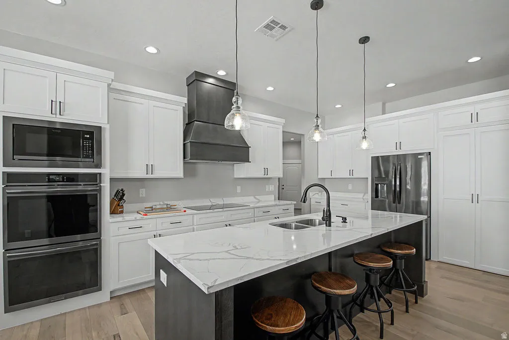 Modern kitchen featuring a large central island with integrated sink, stainless steel built-in appliances, white shaker cabinetry, a dark metal range hood, and light wood-finish flooring