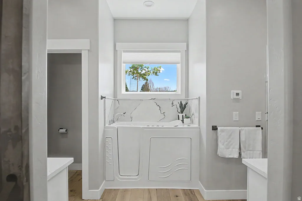 Walk-in tub with white marble-look surround, wood-finish flooring, and a single window with privacy blind