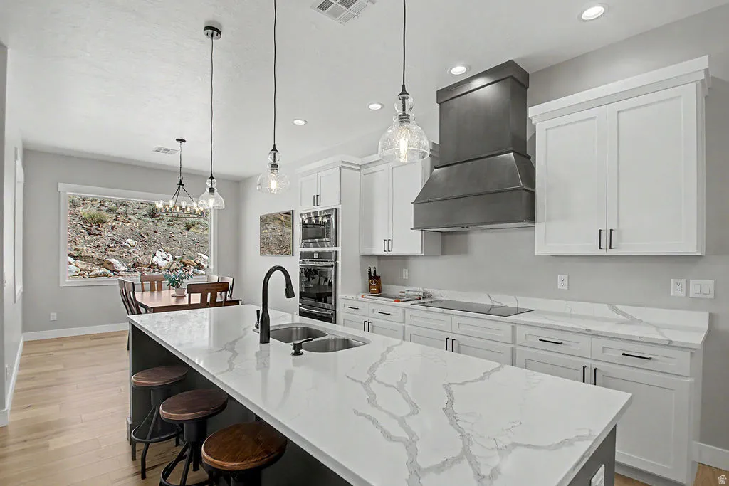 Kitchen featuring an expansive island with a double basin sink, light-toned wood-finish flooring, white shaker-style cabinetry with dark hardware, built-in double ovens, and a custom range hood