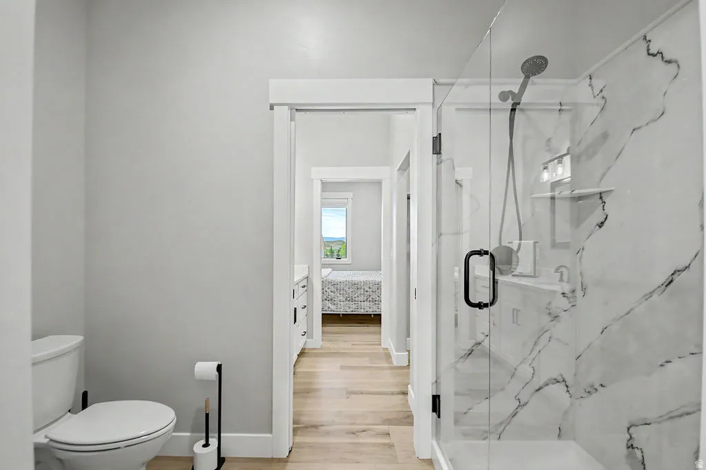 Modern bathroom featuring a glass-enclosed shower with marble-style surround, a white toilet, and wood-finish flooring