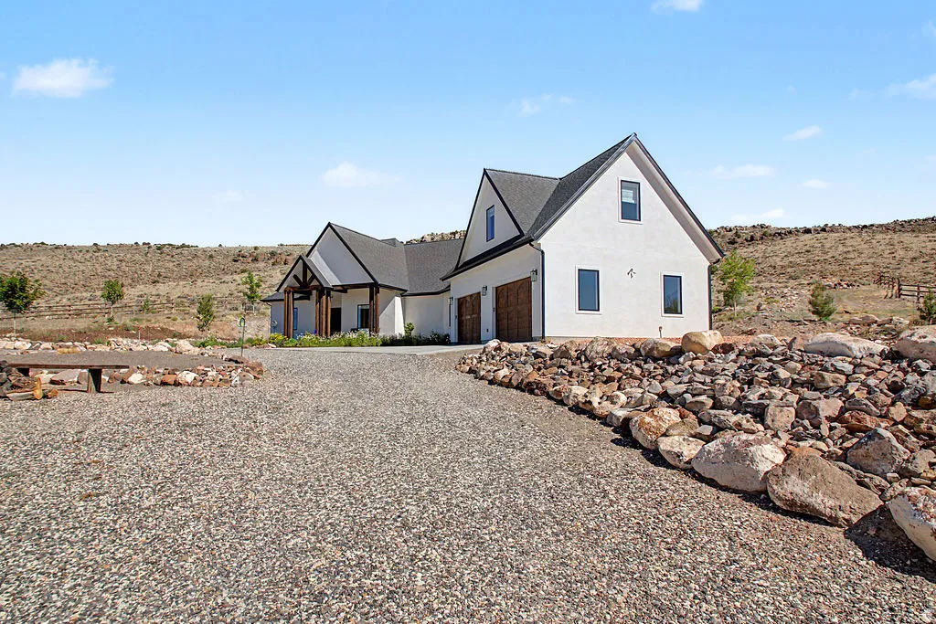 Modern farmhouse exterior with white stucco siding, dark trim, a two-car garage with wood-look doors, a gravel driveway, and a natural rock retaining wall