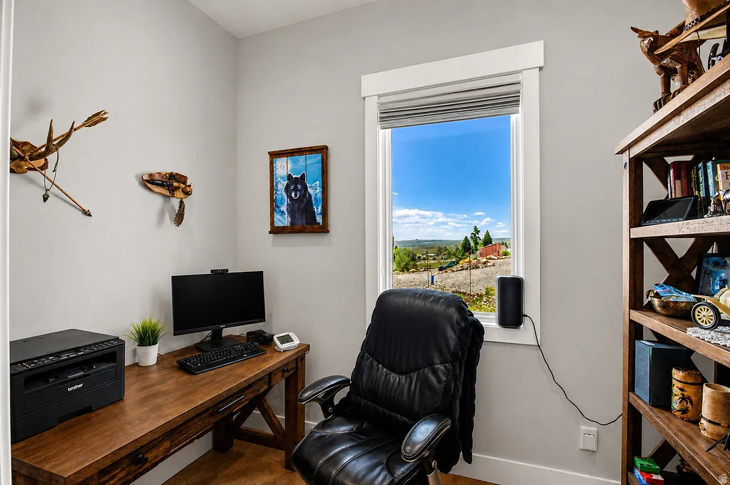 Room with a large window and white trim, featuring a wood-finish desk, wood-finish shelving unit, and light gray wall paint