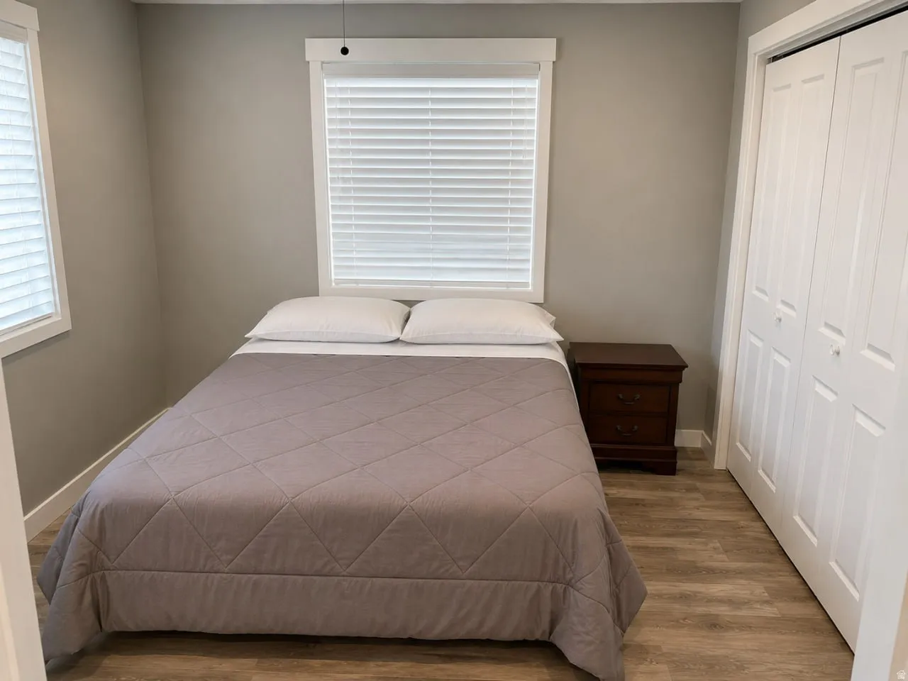 Room featuring neutral gray walls, wood-finish flooring, two windows with white blinds, and a white bi-fold closet door