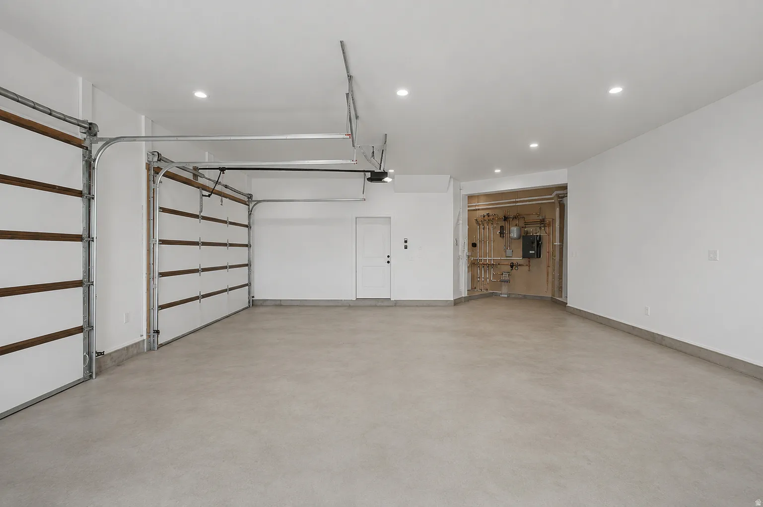 Spacious garage featuring two overhead doors with wood-finish panels, a smooth concrete floor, recessed ceiling lighting, a utility area with exposed piping, and a white interior door