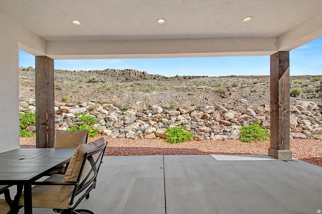 Covered patio featuring recessed lighting, exposed wood-finish support beams, concrete flooring, and a rock-lined perimeter with natural landscaping