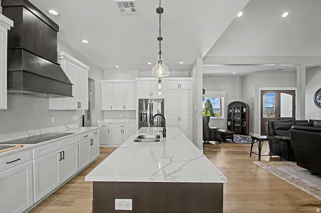 Open-concept kitchen featuring an expansive island with a stainless steel sink, solid surface countertops, light-toned wood-finish flooring, white cabinetry with dark hardware, and a custom dark metal range hood