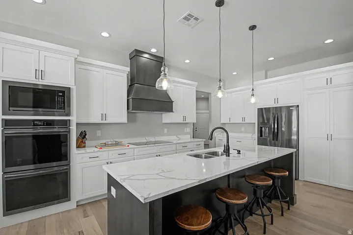 Modern kitchen featuring a large central island with stainless sink, stainless steel built-in appliances, white shaker cabinetry, a dark dark wood range hood, and light Hallmark Orris Maple  flooring