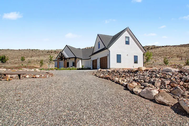 Modern farmhouse exterior with white stucco siding, dark trim, a two-car garage with wood-look doors, a gravel driveway, and a natural rock retaining wall