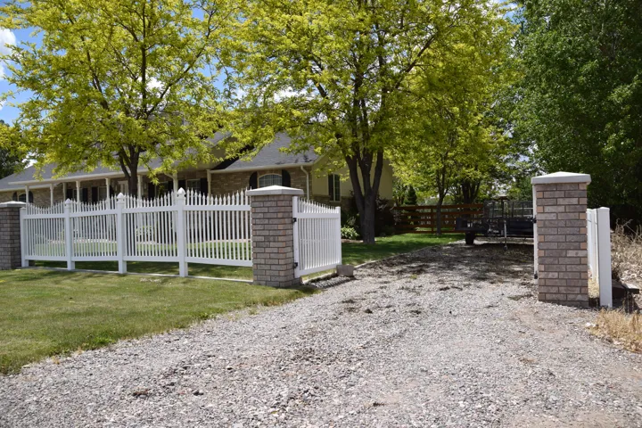 Gravel driveway leading to a white picket fence with brick pillars, surrounding a property with mature trees and a traditional-style home featuring a covered porch