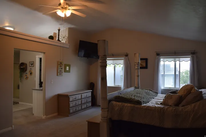Spacious bedroom featuring vaulted ceilings, a ceiling fan with integrated lighting, and two windows providing natural illumination