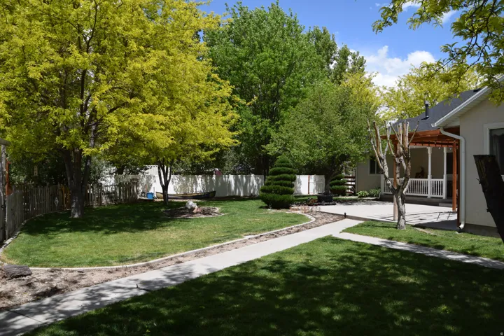 Expansive backyard featuring a manicured lawn, concrete pathways, mature trees, and a covered patio with a white railing and a wood pergola