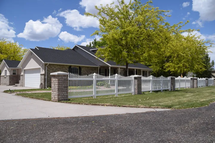 Exterior featuring a white picket fence with brick pillars, a paved driveway, mature trees, and a two-car garage with a white door