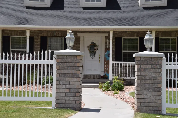 Brick facade residence featuring a covered front porch, white picket fence, and a concrete walkway
