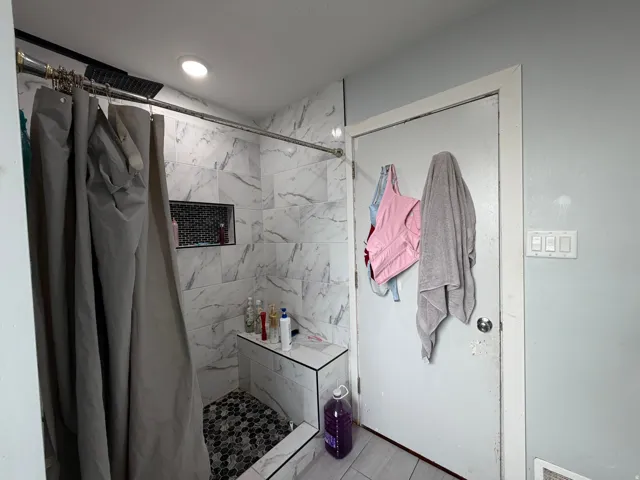 Shower featuring white marble-patterned wall tiles, a built-in bench, a recessed shower niche, a dark pebble-tile floor, and a matte black rainfall showerhead