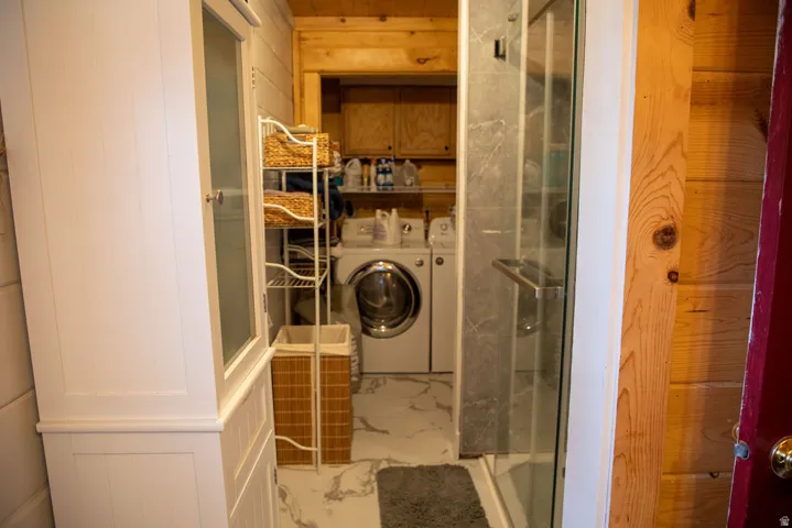 Compact laundry area featuring a front-loading washer and dryer, marble-look flooring, and a glass-enclosed shower with stone-look tile