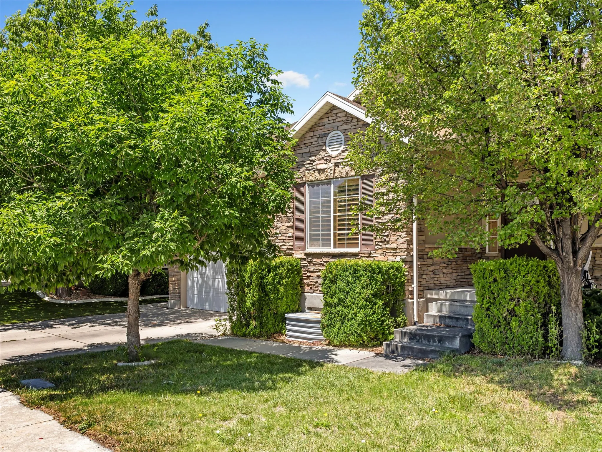 Stone facade residence featuring a two-car garage, mature trees, manicured shrubs, and a concrete driveway