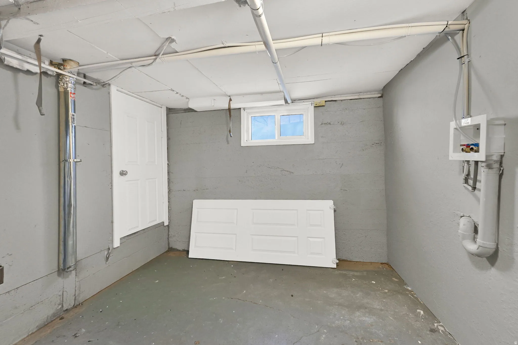 Concrete basement walls with a single window and exposed utility piping