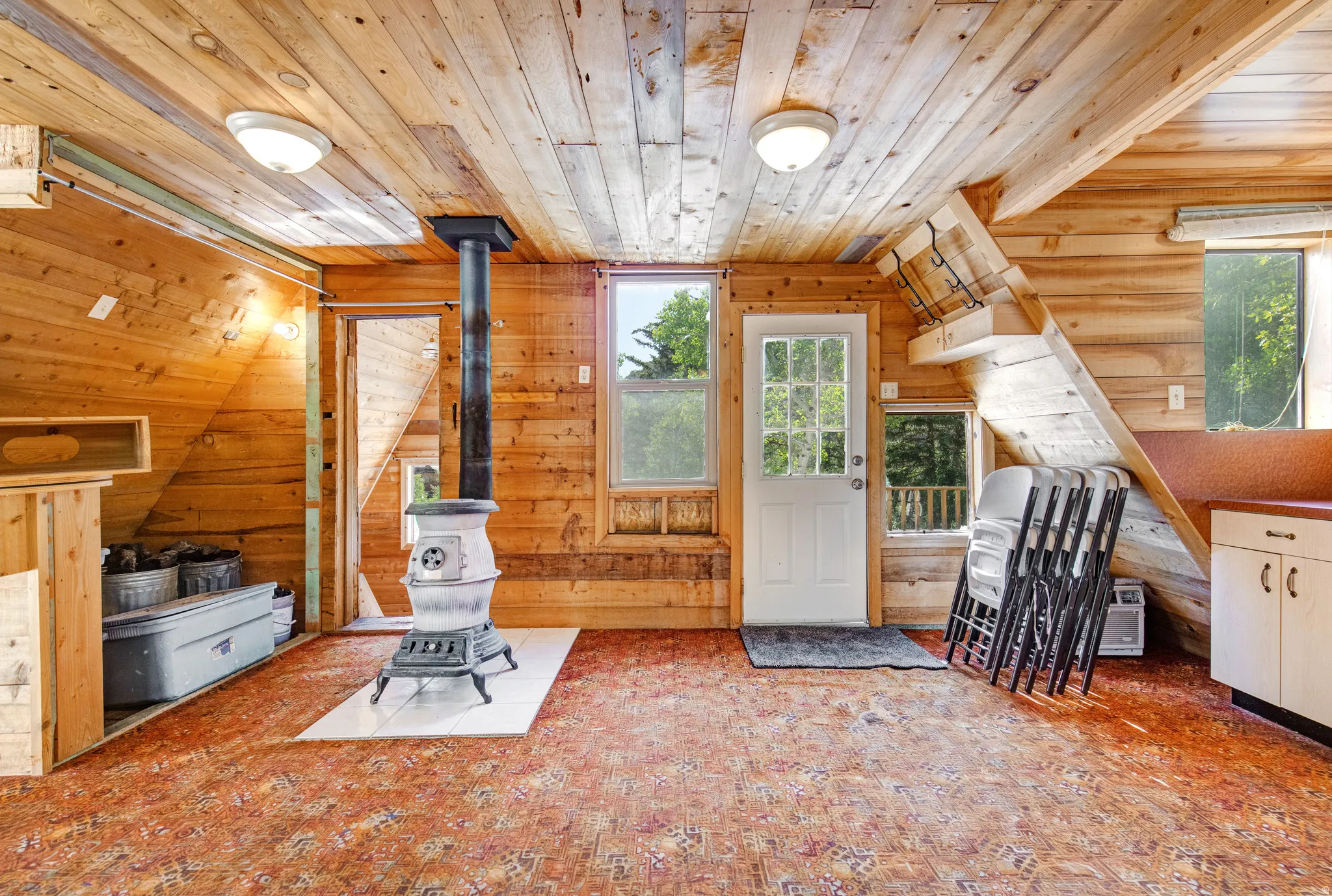 Wood-paneled room featuring a central wood-burning stove on a tile hearth, wood-finish ceiling, and multiple windows