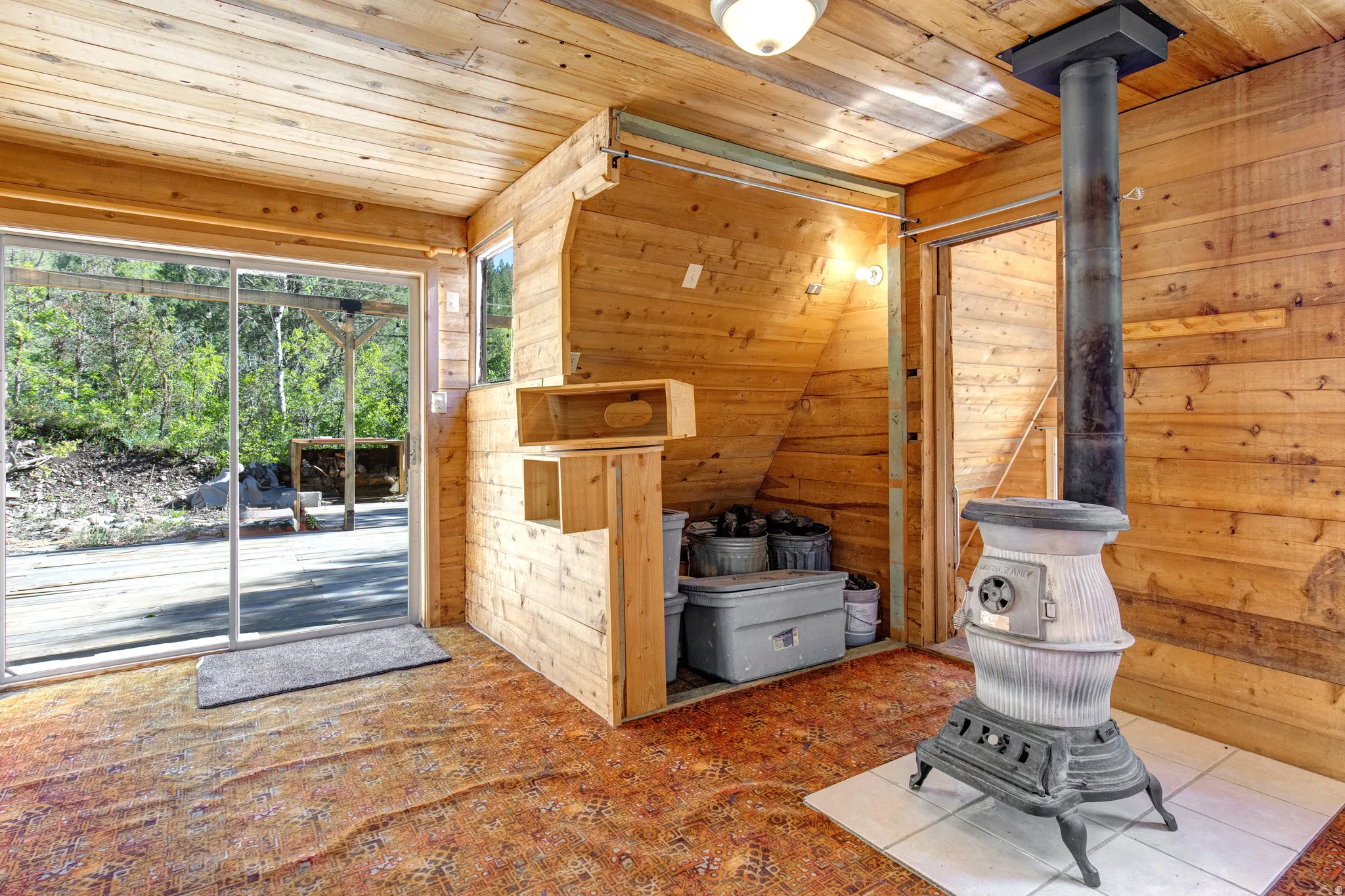Room featuring natural wood plank walls and ceiling, a classic cast iron wood-burning stove on a tile hearth, and a glass sliding door leading to an outdoor area