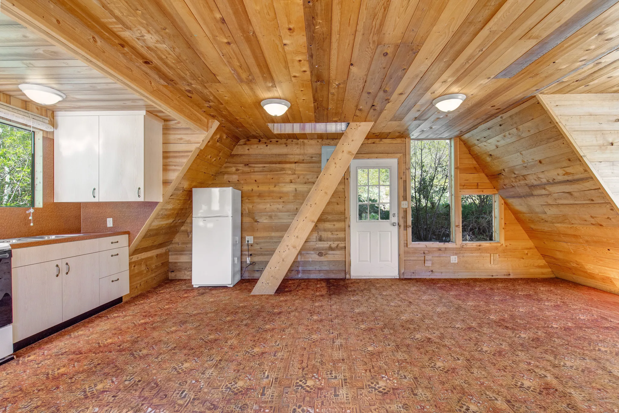 Interior space featuring extensive wood paneling on walls and ceilings, a partial kitchen area with white cabinetry, a white refrigerator, a white door with multiple glass panes, and patterned carpet flooring