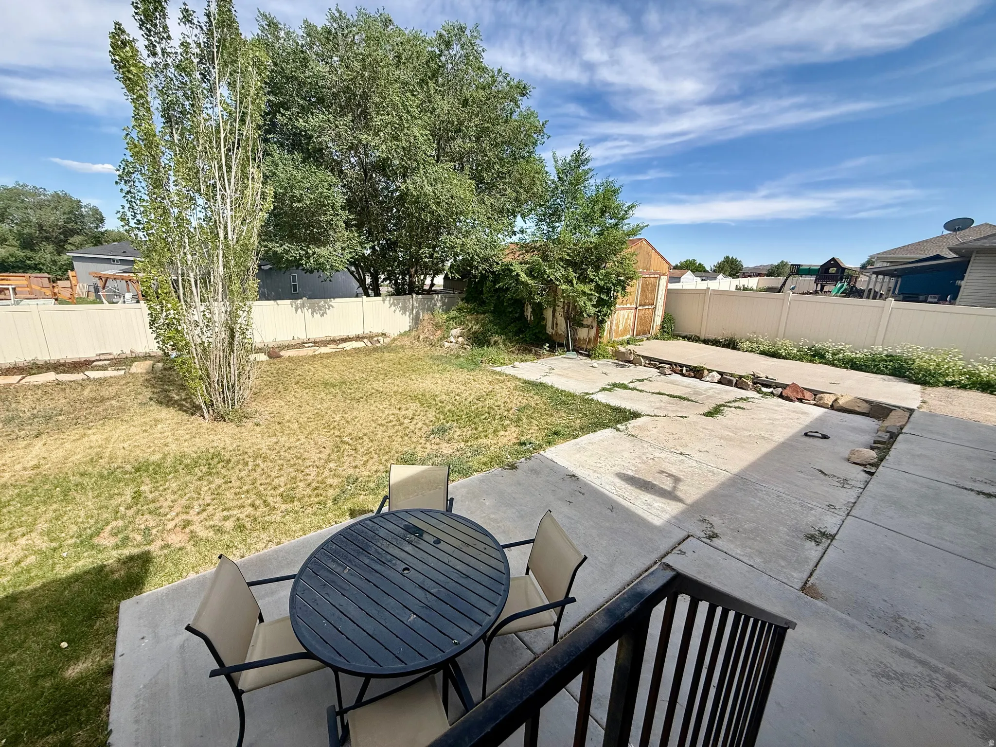 Expansive concrete patio with black railing, opening to a backyard with mature trees and a vinyl privacy fence