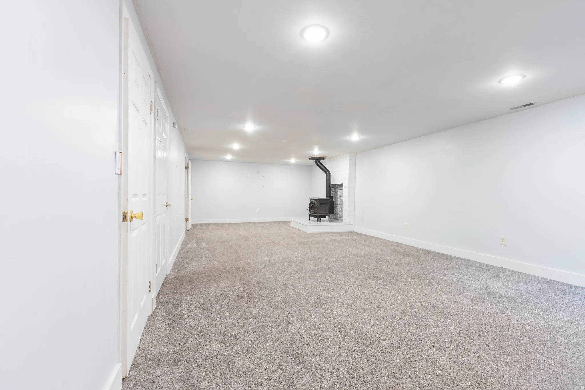 Spacious room featuring light gray carpeting, recessed ceiling lighting, and a black wood-burning stove with a stone surround
