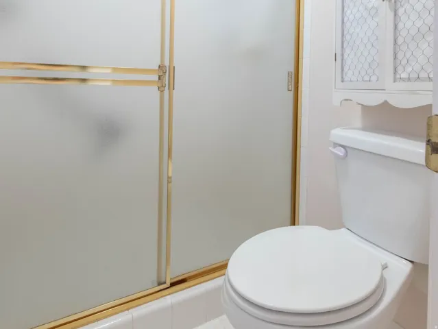 Bathroom featuring a white toilet, white tile flooring, and a shower with frosted glass and a gold-finish frame