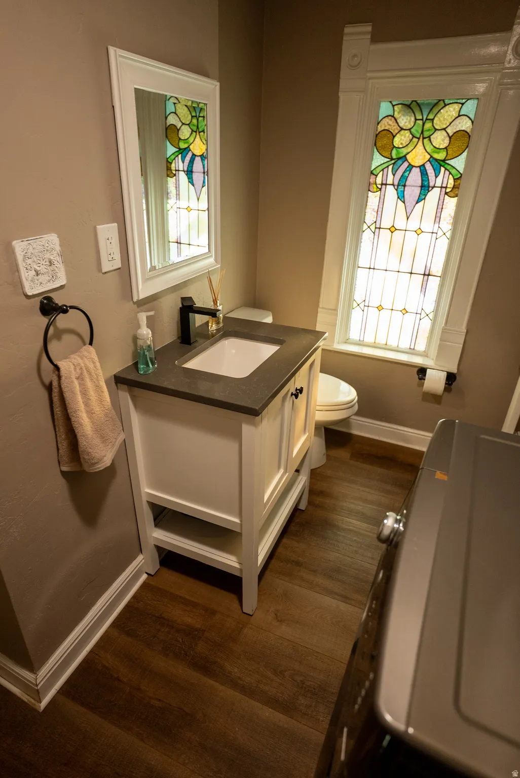 Bathroom featuring a stained-glass window, white vanity with a dark countertop, white framed mirror, matte black faucet, and wood-finish flooring