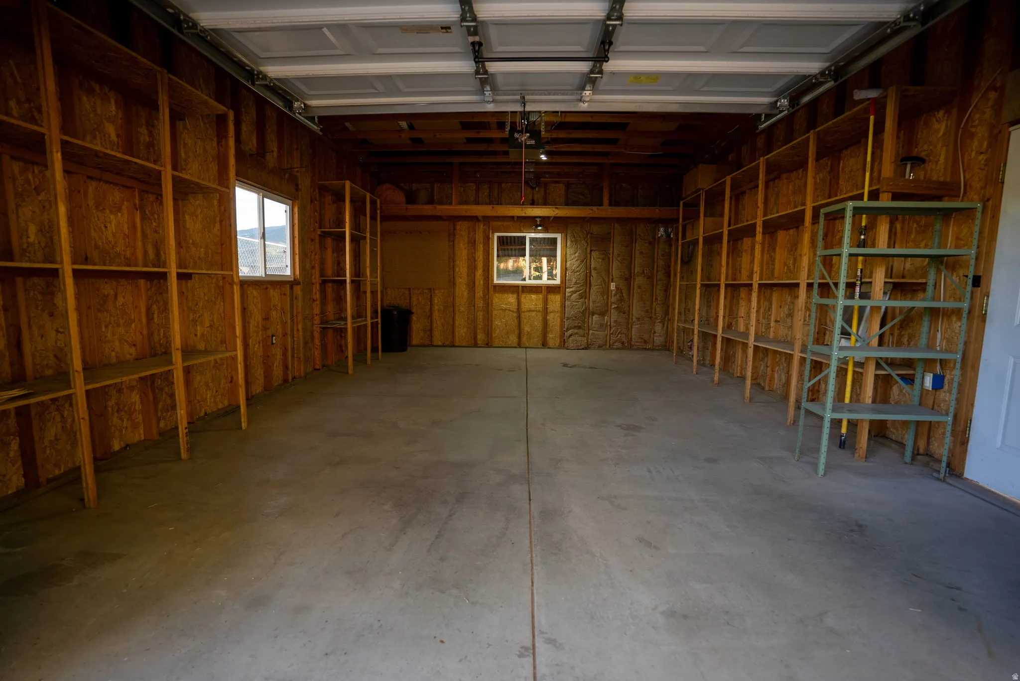 Spacious garage featuring a concrete floor, exposed wood-finish walls, multiple built-in shelving units, two windows, and a panel garage door
