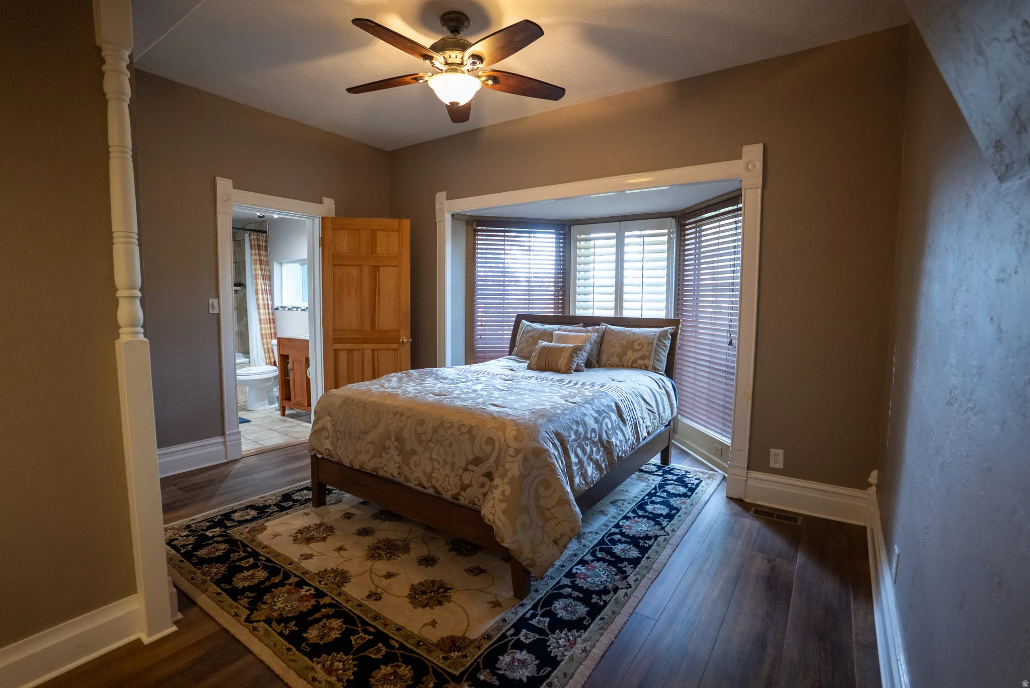 Bedroom featuring wood-finish flooring, a ceiling fan with integrated lighting, a bay window with horizontal blinds, and an entryway to an attached bathroom